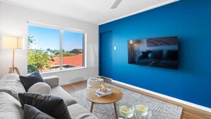 A modern living room with a large window, a ceiling fan, a flat-screen TV, and a cozy seating area. - Azure Walk - Short Walk to the Beach (Scarborough)