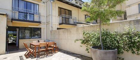 A courtyard with a wooden table and chairs, a large potted plant, and a building with balconies.