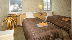 A hotel room with two beds, a desk, a chair, a lamp, and a window with a view of buildings and mountains.