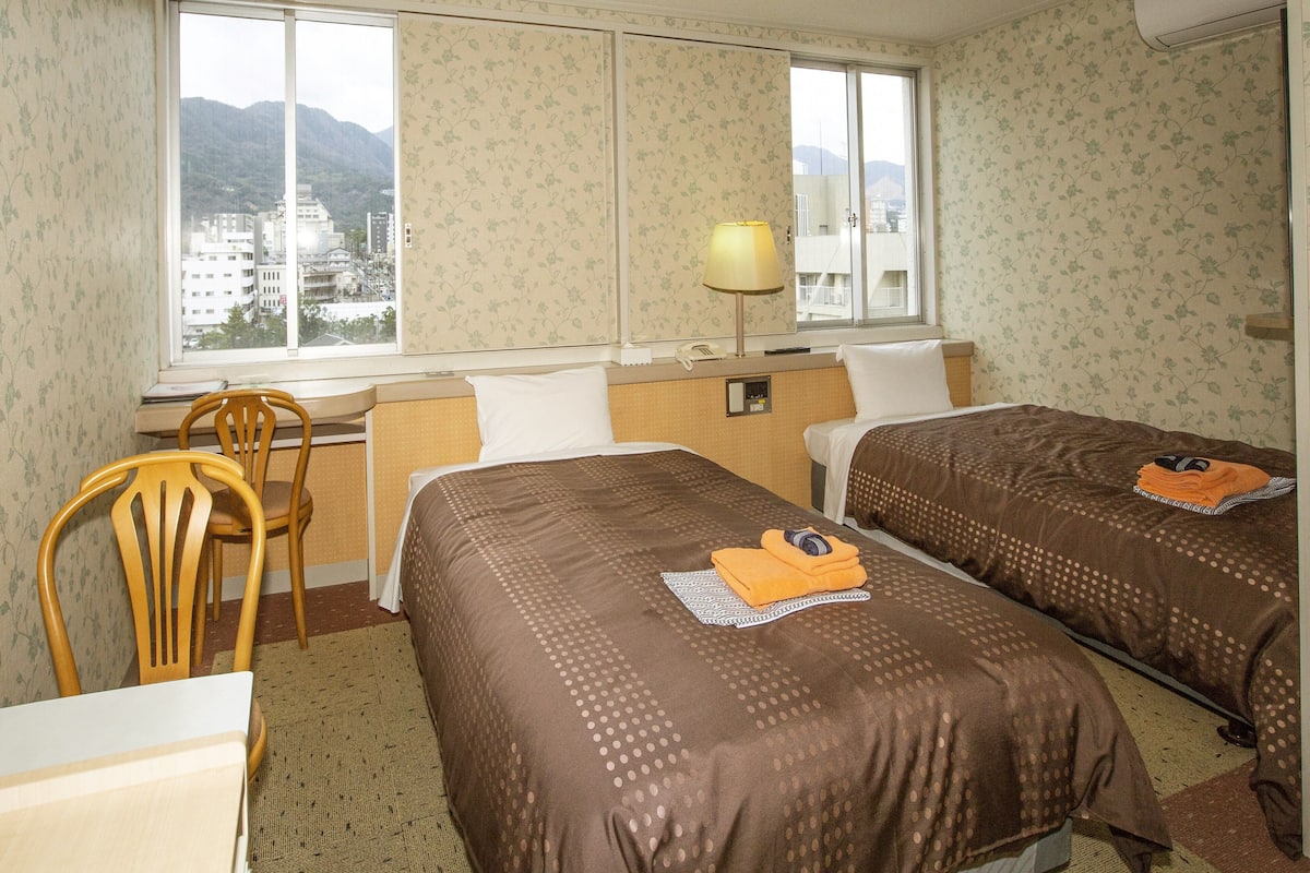 A hotel room with two beds, a desk, a chair, a lamp, and a window with a view of buildings and mountains.