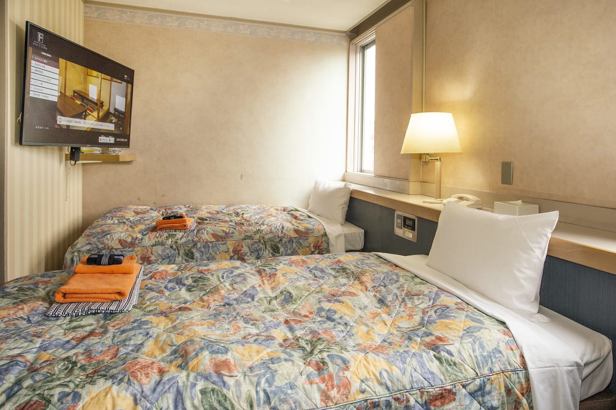 A hotel room with two beds, a TV, a lamp, and a window.