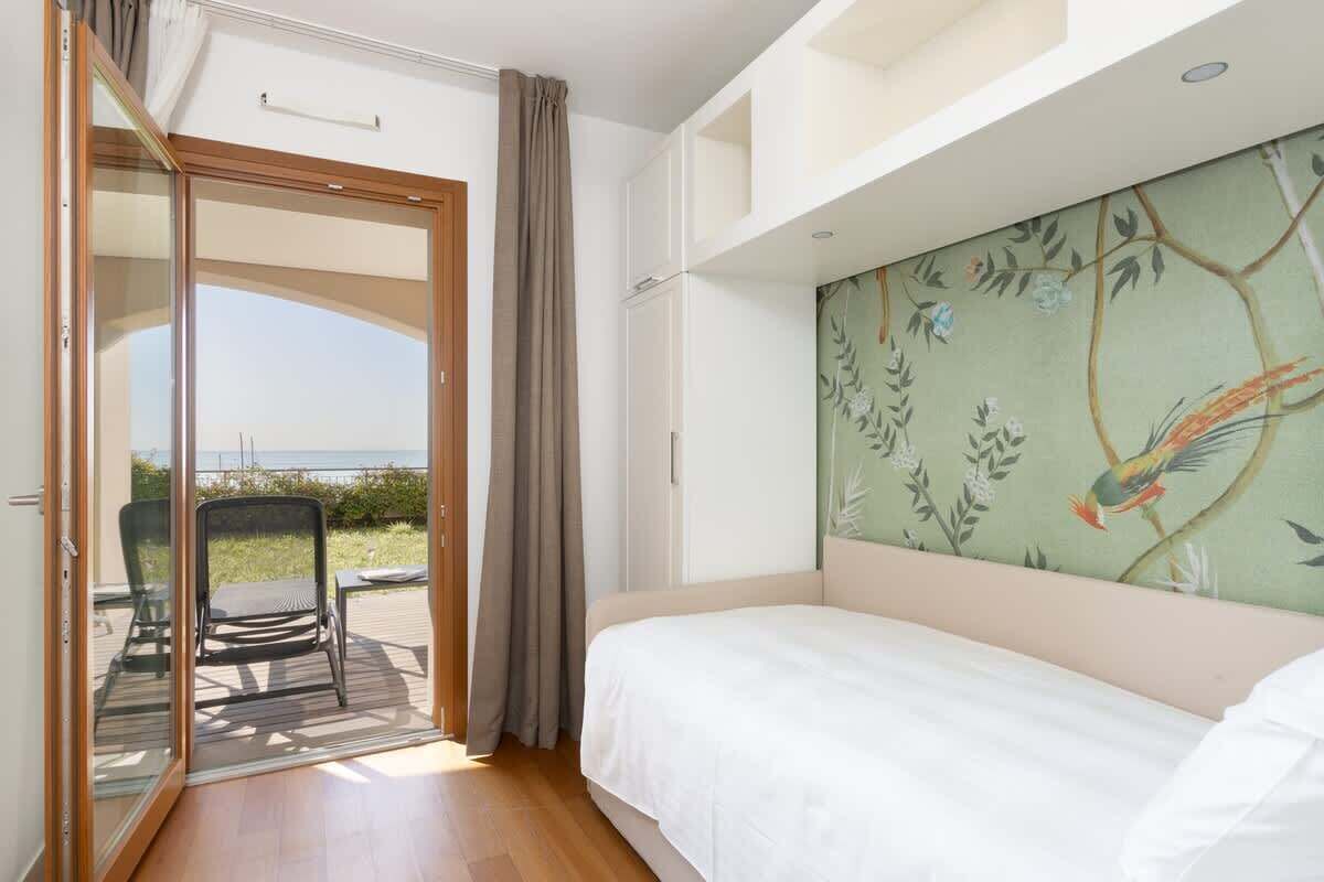 Un dormitorio con una cama, un ventanal corredero que da a un área exterior con una silla y una pared decorada con un mural de temática natural.