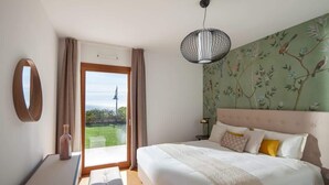 Ein Schlafzimmer mit Bett, Nachttisch, Lampe, Fenster mit Blick und einer Wand mit Blumenmuster.