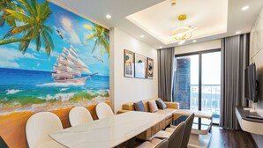 A modern living room with a large painting of a sailboat on the ocean, a sofa, a dining table with chairs, and a TV on the wall. - X-Stay Luxe (Hai Phong)