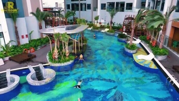 A large indoor pool with multiple levels and water slides, surrounded by palm trees and greenery. - X-Stay Luxe (Hai Phong)