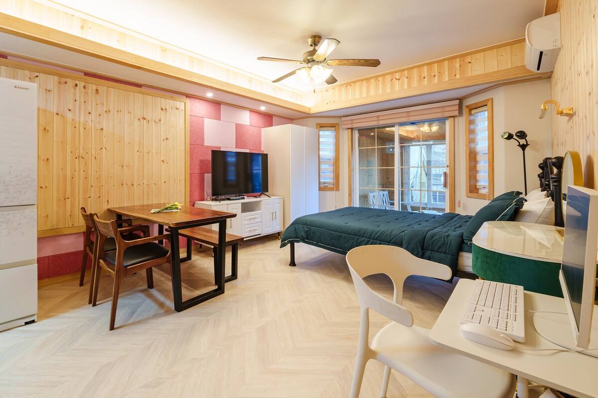 Une pièce de taille modeste comprenant un lit, un bureau avec un ordinateur, une table à manger avec des chaises, un téléviseur et un réfrigérateur.