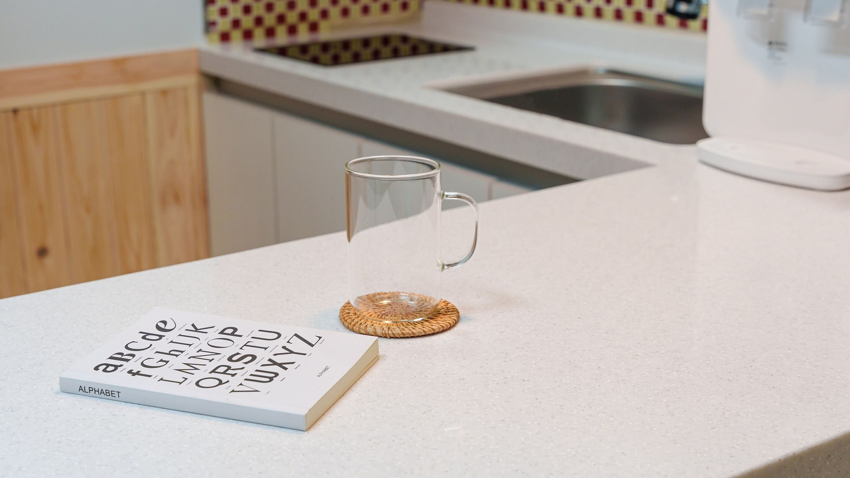 A kitchen counter with a book titled 'alphabet' and a glass mug on a woven coaster.