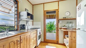 Una cocina con armarios de madera, estufa, microondas y fregadero. Se ve una puerta que da al exterior con vista a vegetación.