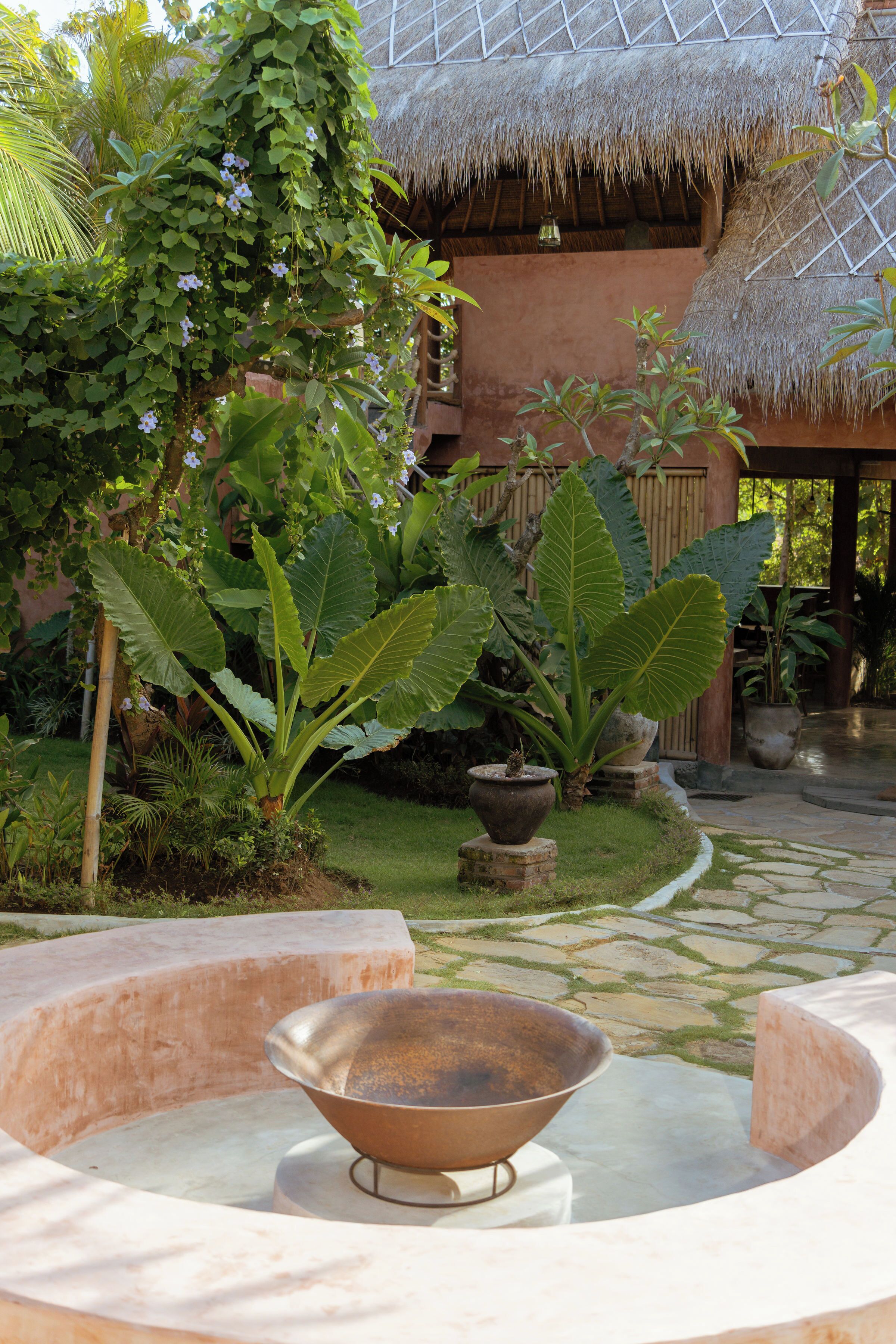 Un patio con una estructura de techo de paja, plantas grandes en macetas y un cuenco metálico en el centro.
