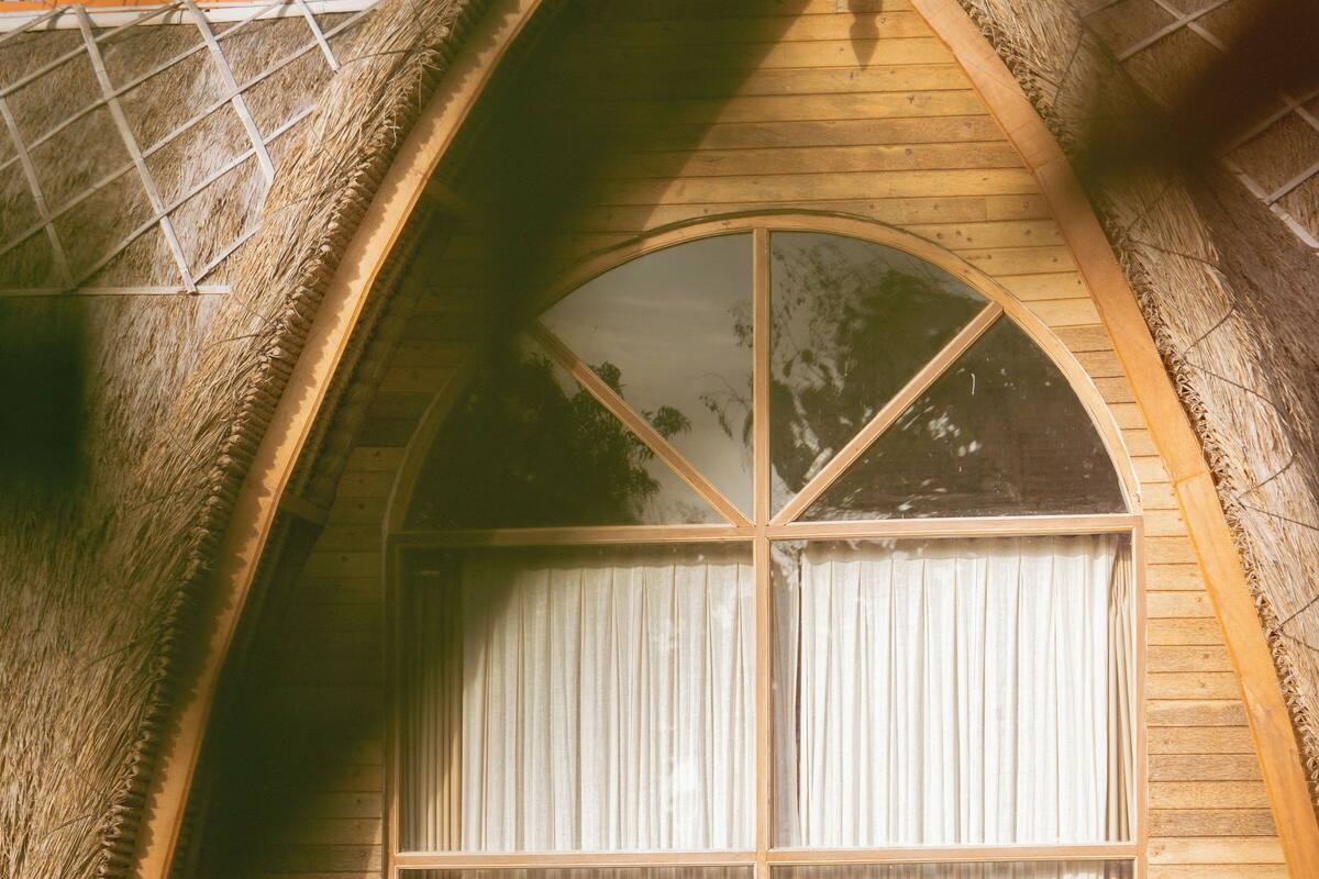 Un bâtiment à toit de chaume doté d’une grande fenêtre et de rideaux.