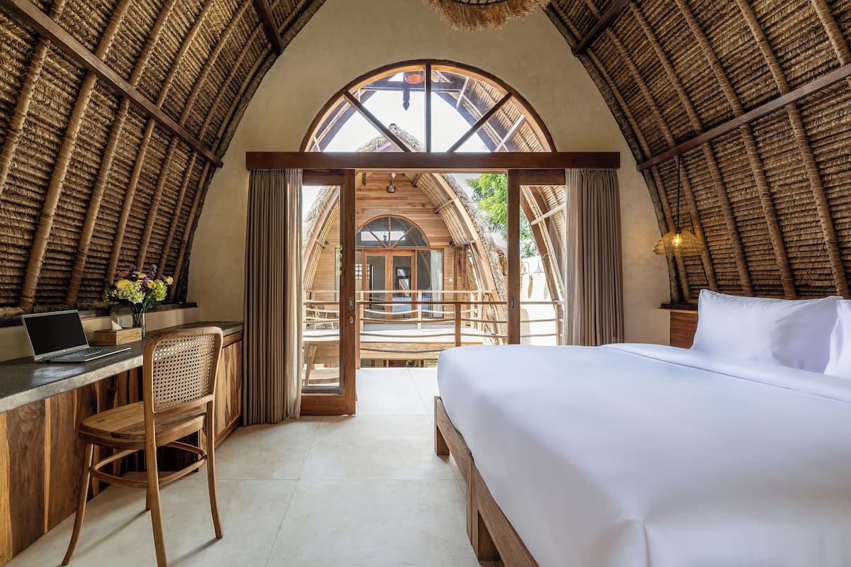 Une pièce comprenant un lit, un bureau et une chaise, avec une grande fenêtre en arc de cercle et des poutres en bois apparentes au plafond.