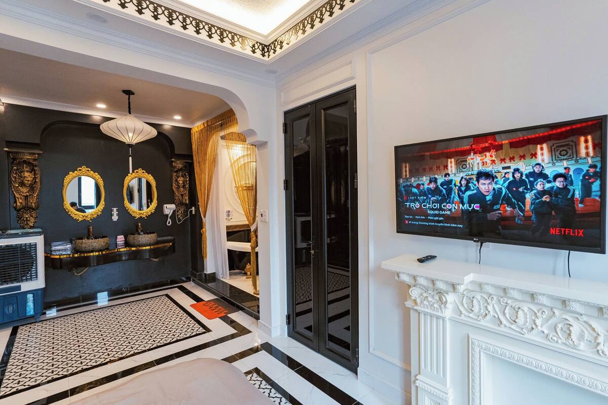 A luxurious lobby with a black and white tiled floor, a large mirror with golden frames, a chandelier, and a television displaying a Netflix show.