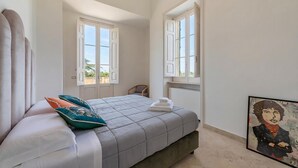 Une chambre à coucher avec un lit, deux fenêtres, une chaise et un tableau accroché au mur.