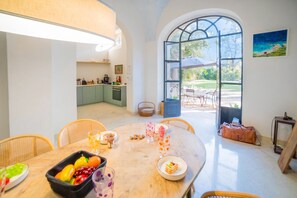 A dining area with a table set for two, a kitchen in the background, and a large window with a view of the outdoors.