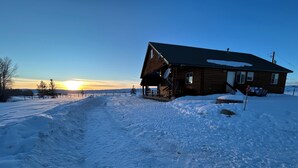 Una cabaña de madera con porche, rodeada de nieve y árboles, con un cielo despejado al fondo.