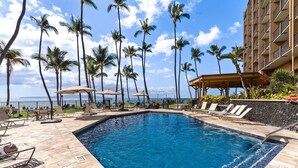 A swimming pool with lounge chairs and umbrellas, surrounded by palm trees and a view of the ocean.