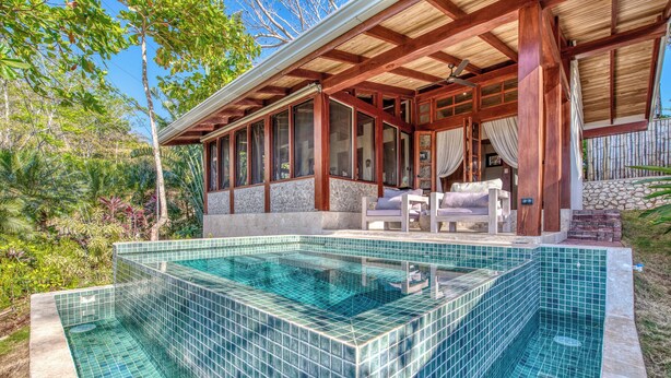 Une piscine avec une terrasse carrelée, un espace salon couvert comprenant un canapé et des fauteuils, et un mur en pierre.