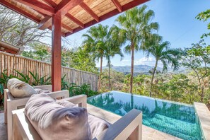A pool with a view of palm trees and mountains, wooden lounge chairs, and a bamboo fence.
