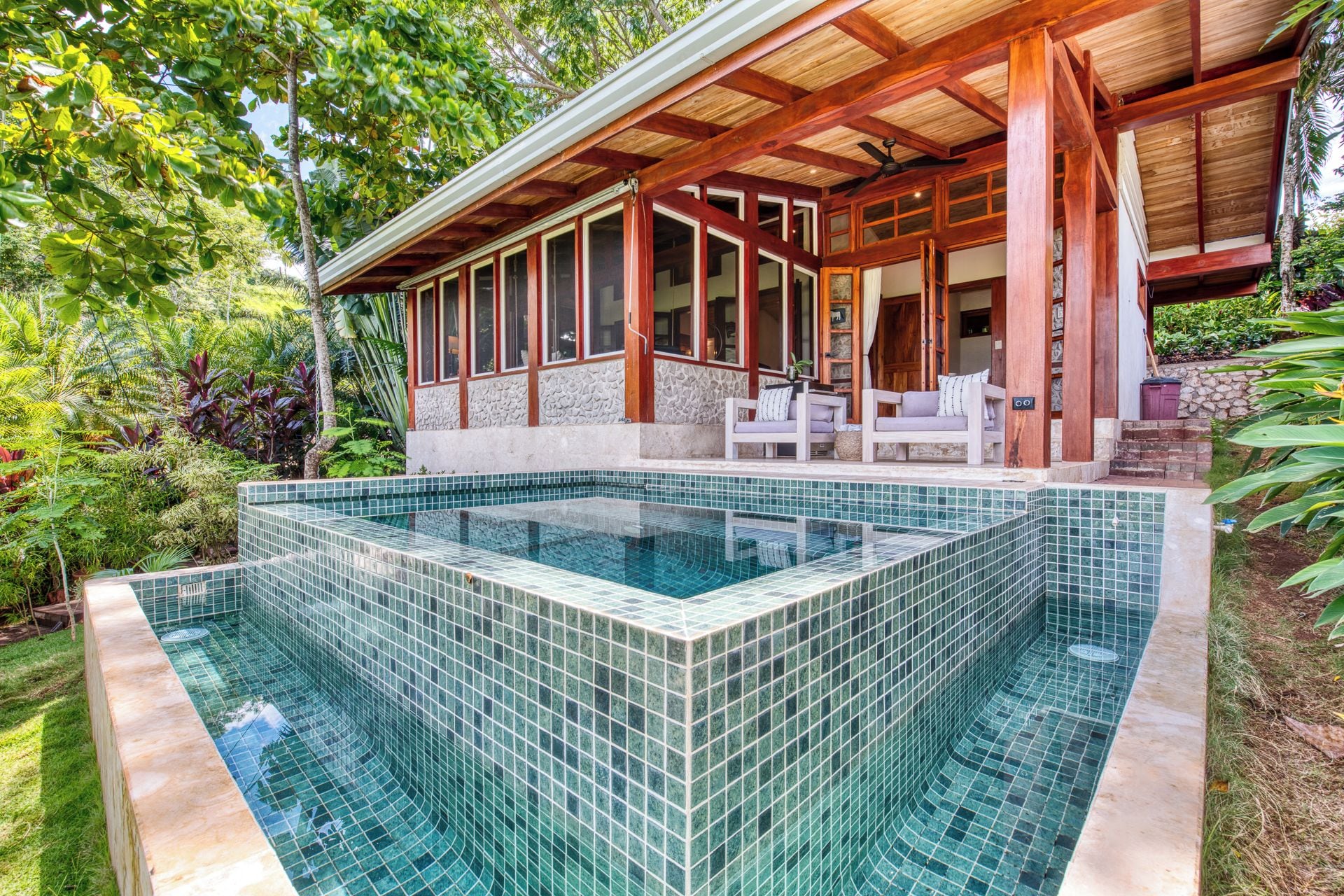 A swimming pool with a tiled edge, a covered patio with seating, and a stone wall.