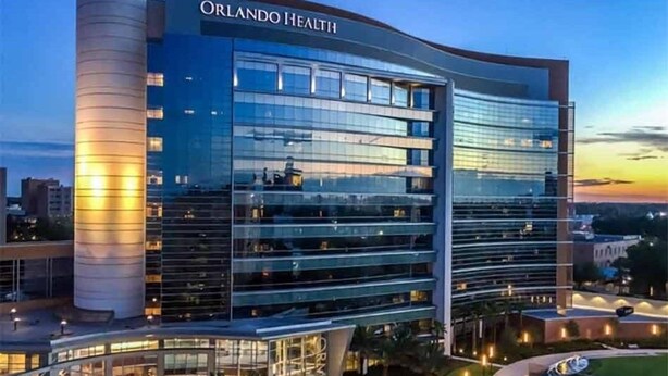 Un bâtiment moderne à plusieurs étages, doté d’une façade en verre, appelé Orlando Health, et qui se distingue par une structure cylindrique imposante située sur le côté gauche.