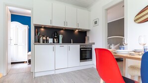 A modern kitchen with white cabinets, a red chair, and a dining table set for two. - Ferienwohnung in Vollerwiek Direkt am Nordseedeich (Vollerwiek)