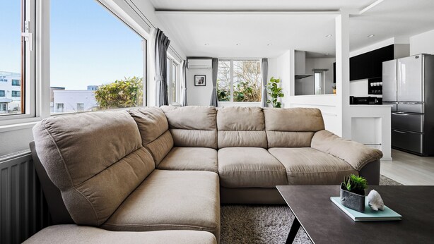 Un salon moderne avec un canapé d’angle, une petite table agrémentée d’une plante et une vue sur la ville grâce à de grandes fenêtres.