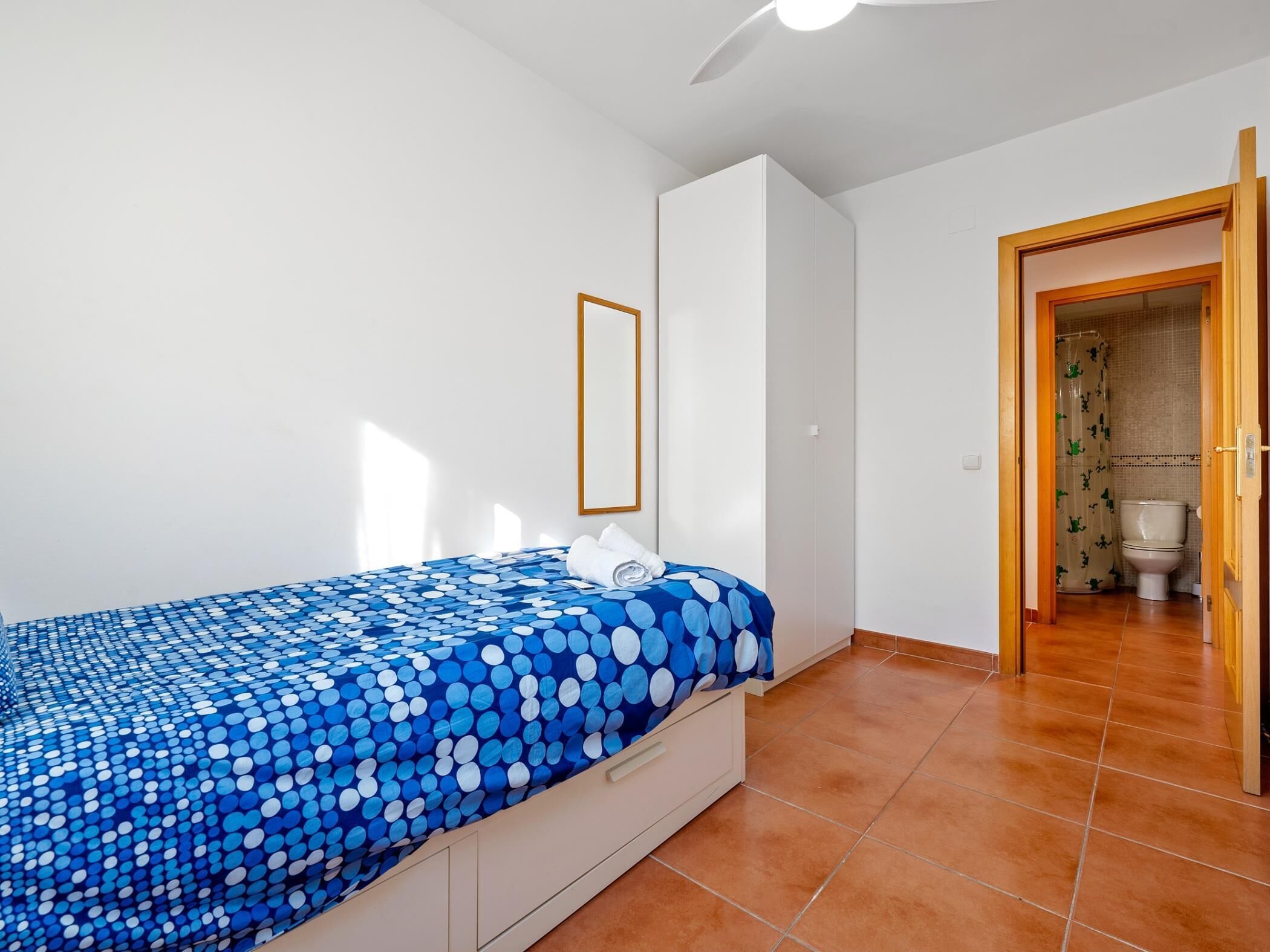 Une chambre à coucher avec un lit, un ventilateur de plafond, une armoire blanche et une salle de bain visible à travers une porte ouverte.