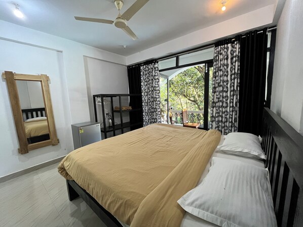 A bedroom with a bed, a ceiling fan, a small desk, a mirror, and a window with curtains. - Hotel God's Gift (Morjim)