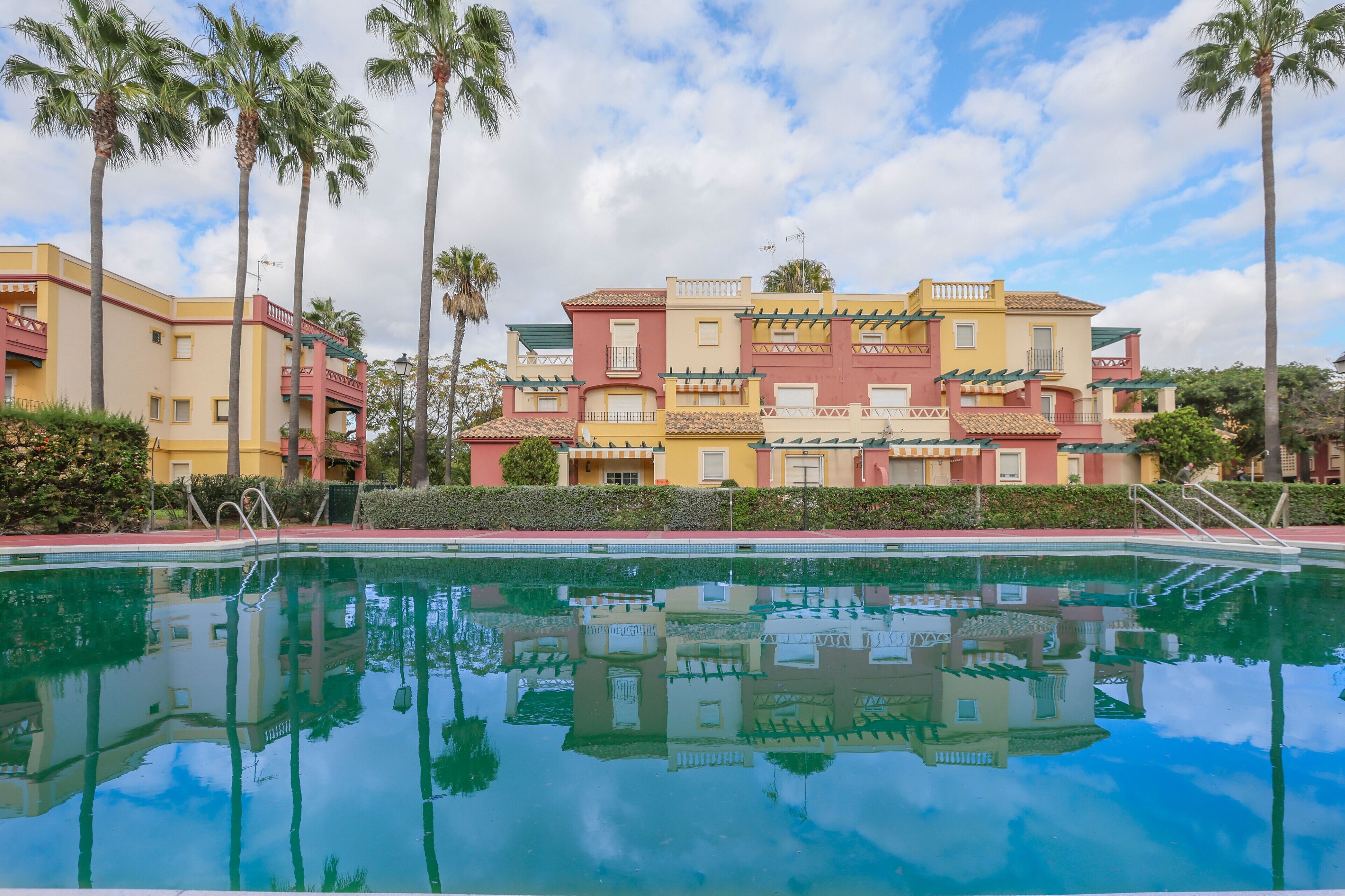 A pool with a reflection of a multi-story building with balconies and palm trees.