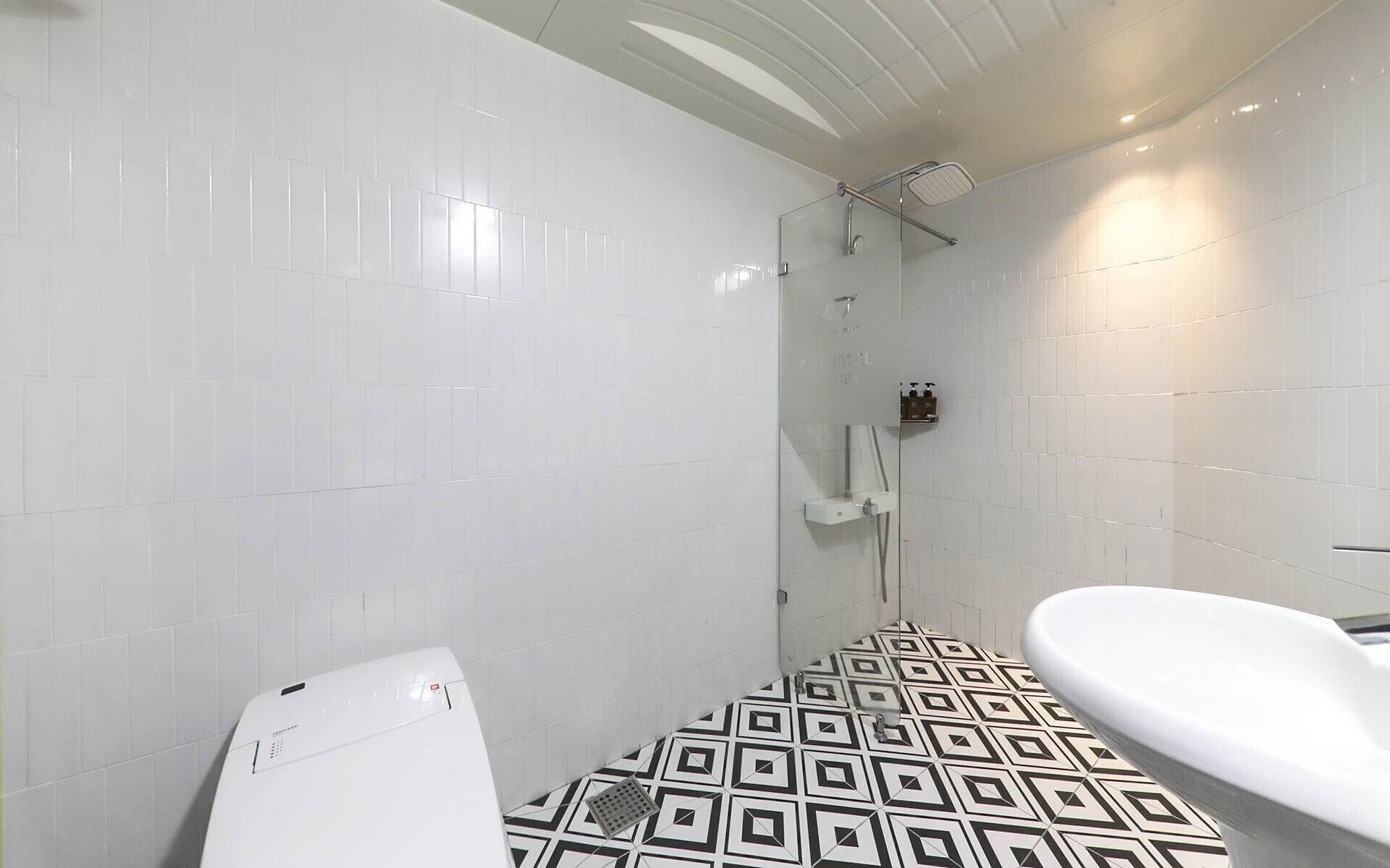 Ein Badezimmer mit weißen Fliesen, einem Boden mit geometrischem Muster, einer Dusche mit Glasabtrennung und einem weißen Waschbecken.