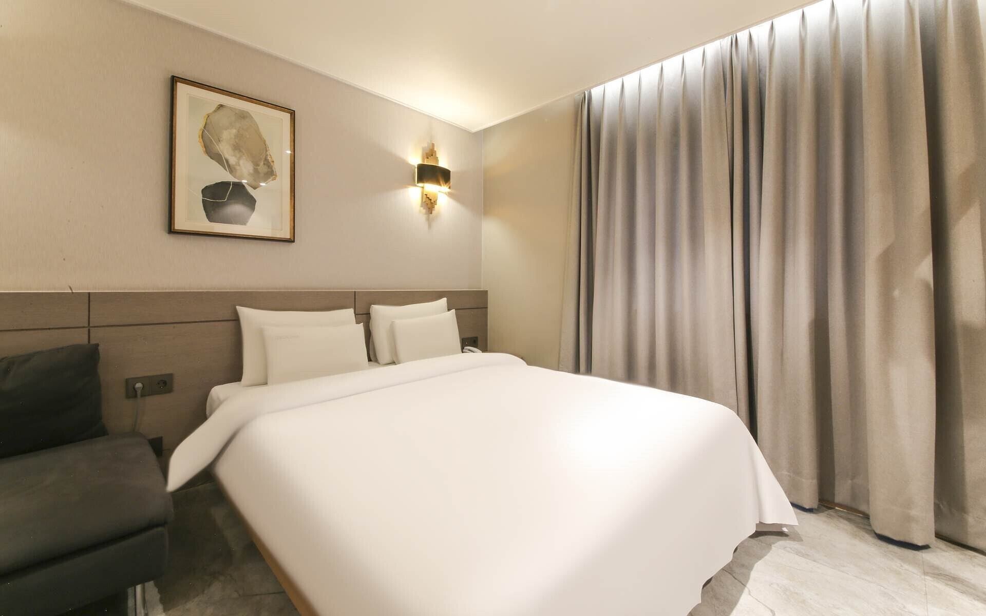 Una habitación de hotel con una cama grande, un cabecero, una lámpara de pared, una obra de arte enmarcada y una ventana con cortinas.