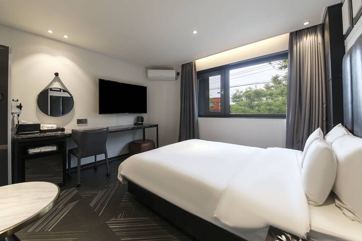 A hotel room with a large bed, a desk with a chair, a flat-screen TV, and a window with curtains.