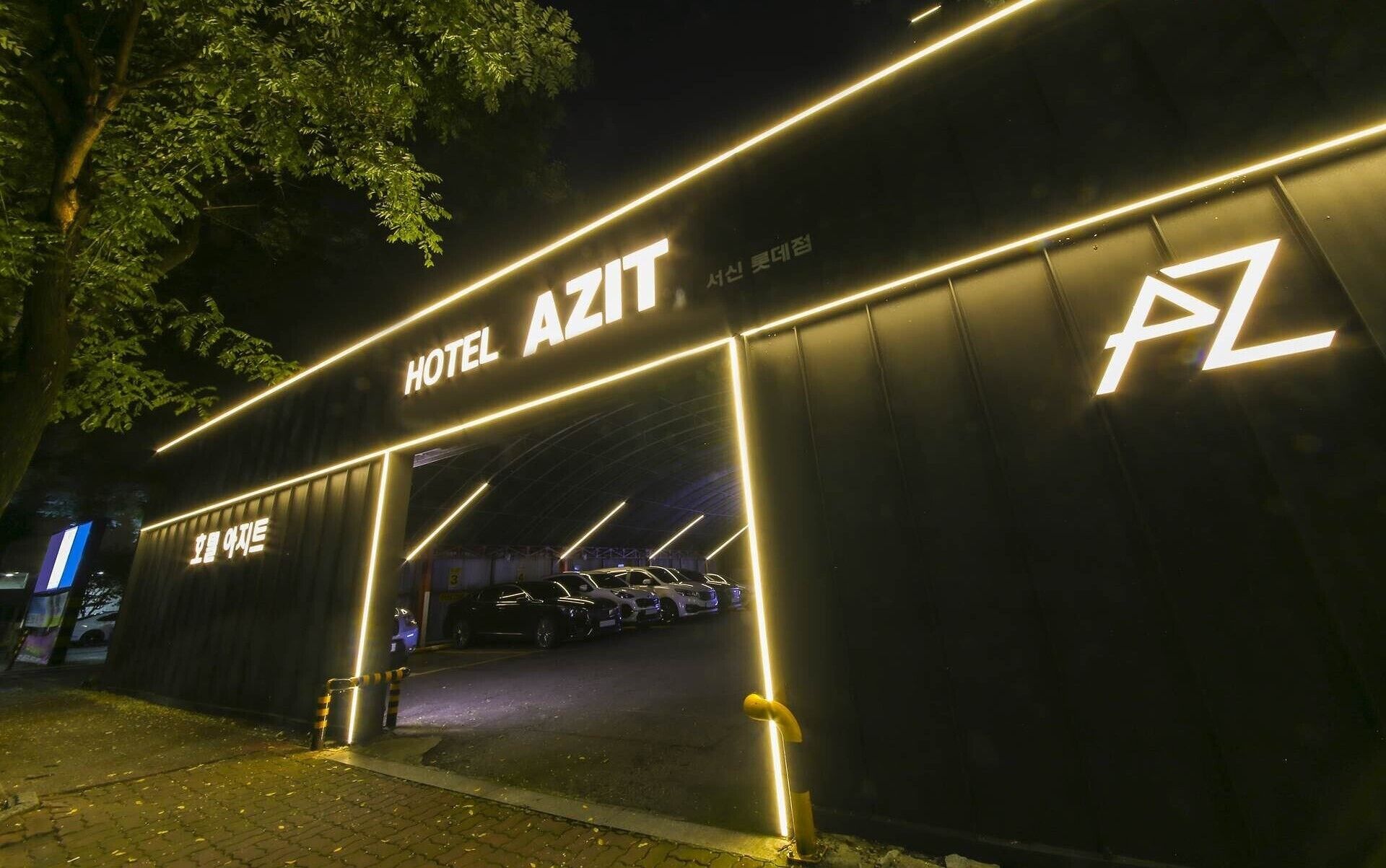 A hotel entrance with illuminated signage reading "HOTEL AZIT" and a parking area with several cars.