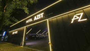 A hotel entrance with illuminated signage reading "HOTEL AZIT" and a parking area with several cars.