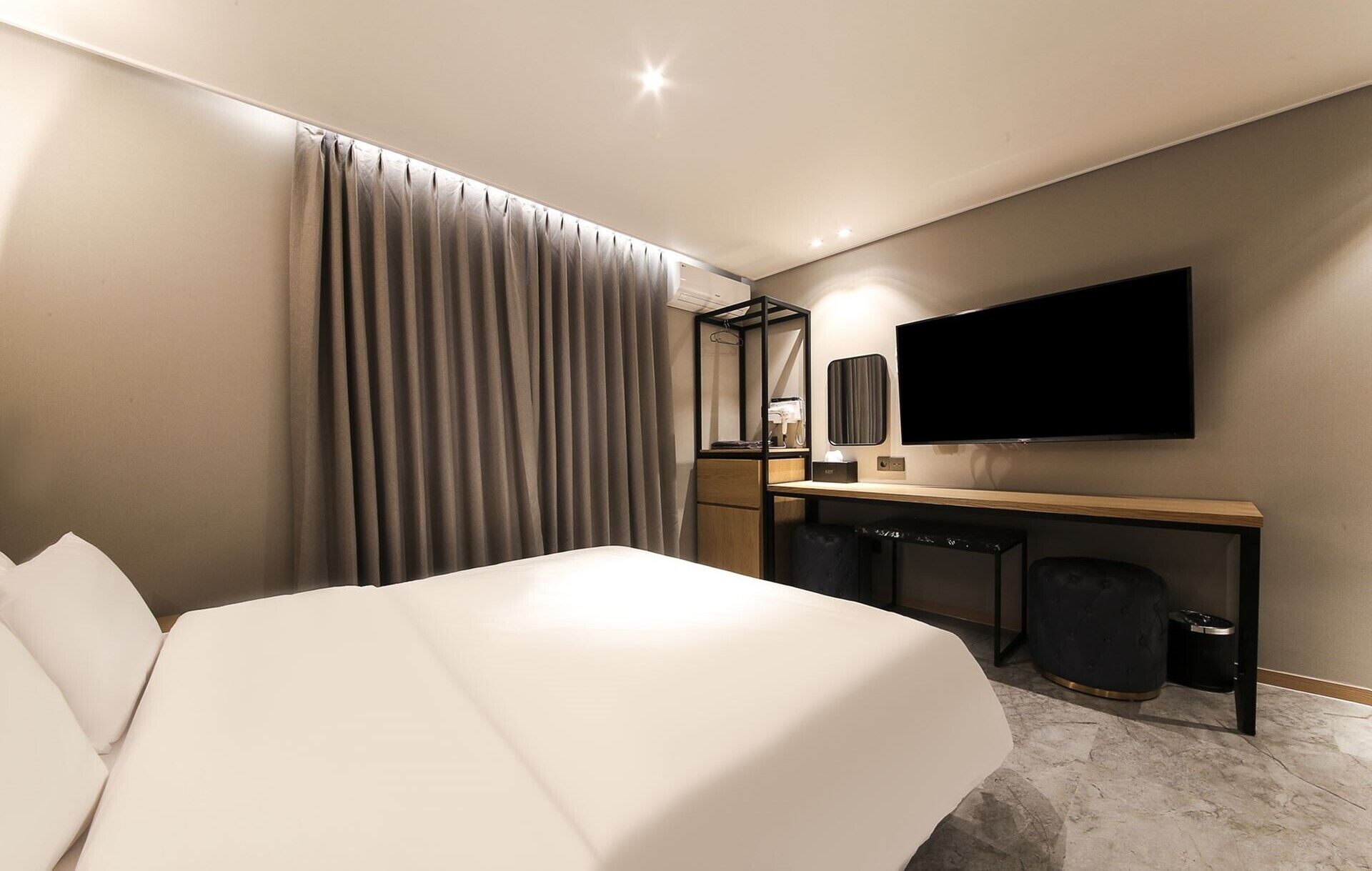 A modern hotel room with a large bed, a desk with a flat-screen TV, a mirror, and a trash bin.