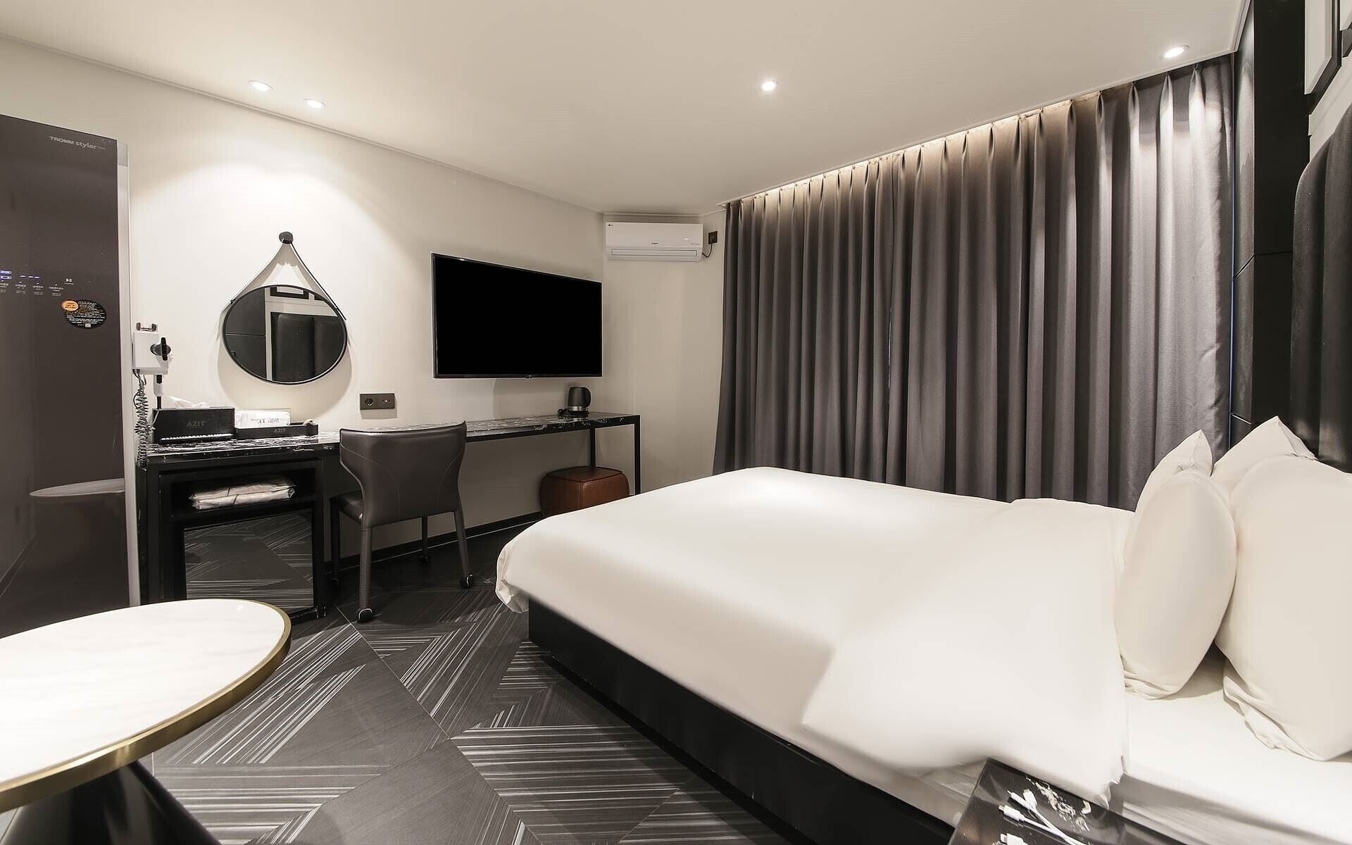 A modern hotel room with a large bed, a desk with a chair, a round table, a flat-screen TV, and a large window with curtains.