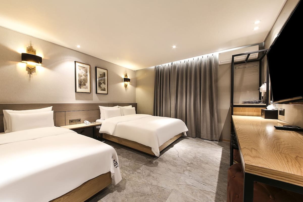 A hotel room with two beds, a desk, a chair, a TV, and a window with curtains.