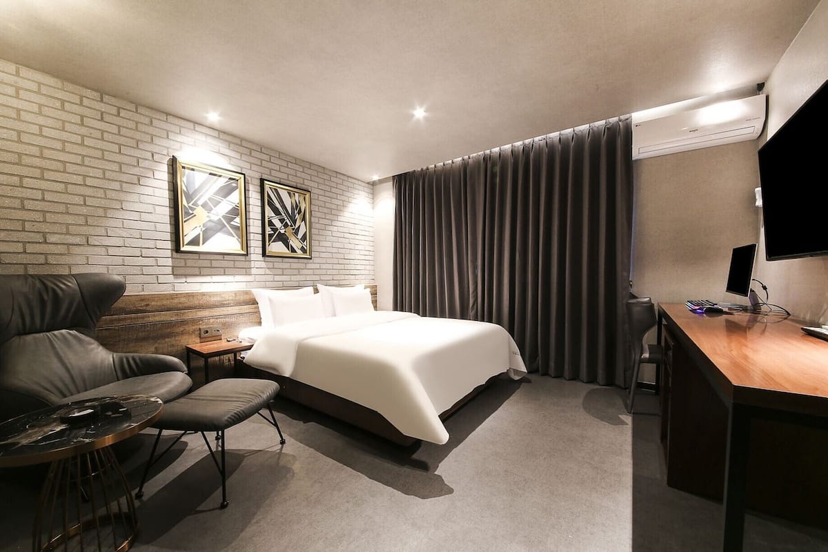 A modern hotel room with a brick wall, a large bed, a desk with a computer, and a seating area with a chair and a small table.