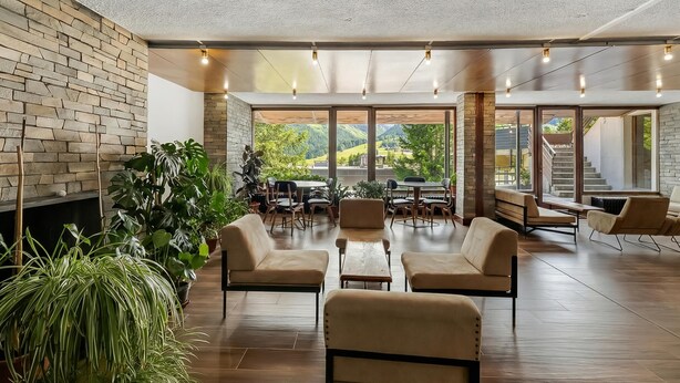 Un espace intérieur vaste et lumineux, doté d’une cheminée, de plusieurs coins salon et de grandes fenêtres offrant une vue sur la verdure et un escalier.