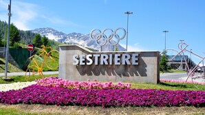 Une pancarte portant l’inscription « SESTRIERE » et les anneaux olympiques, entourée de fleurs et avec une chaîne de montagnes en arrière-plan.