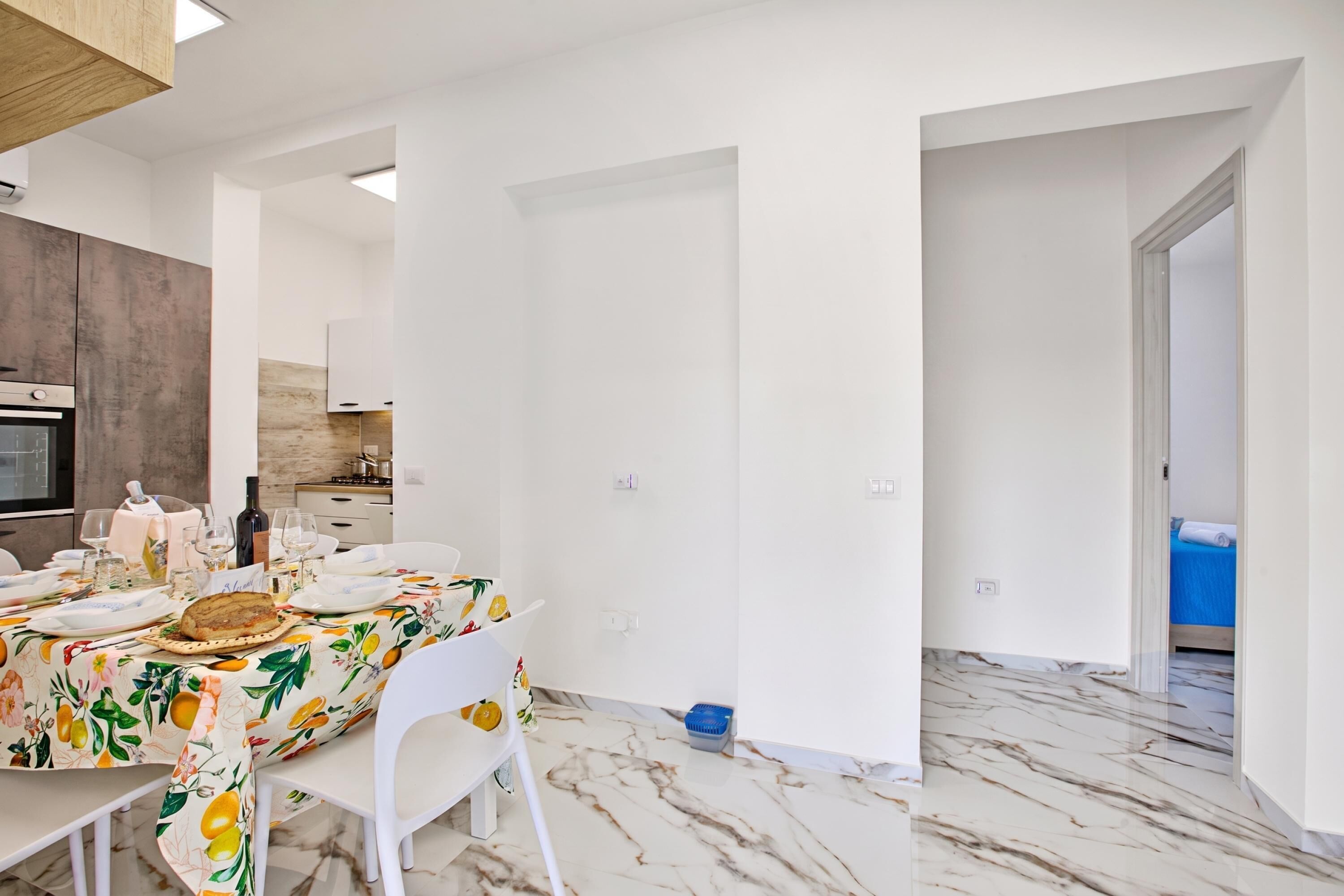 Una cucina moderna con mobili bianchi, un tavolo da pranzo apparecchiato con piatti e bicchieri, e un pavimento in marmo.