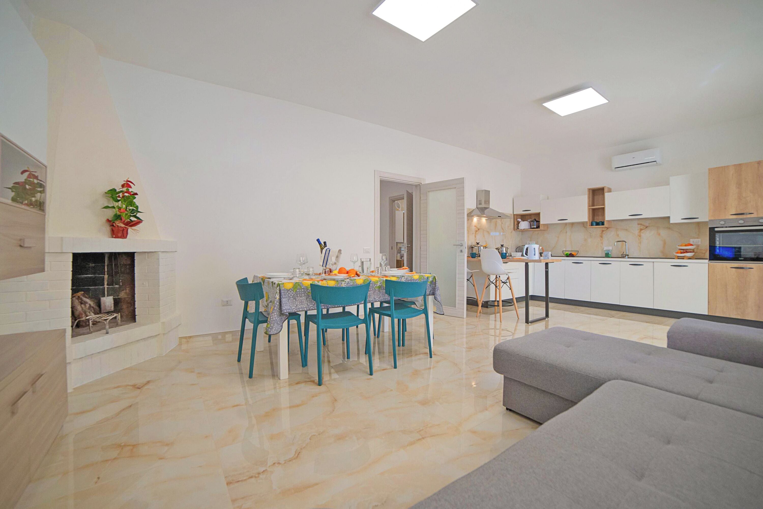 Uma sala de estar moderna com um sofá cinzento, um espaço de refeições com mesa e cadeiras, e uma cozinha com armários brancos e forno embutido.