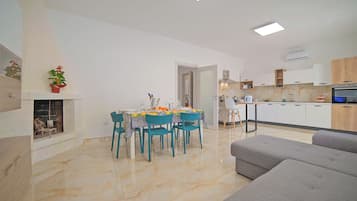 Uma sala de estar moderna com um sofá cinzento, um espaço de refeições com mesa e cadeiras, e uma cozinha com armários brancos e forno embutido.