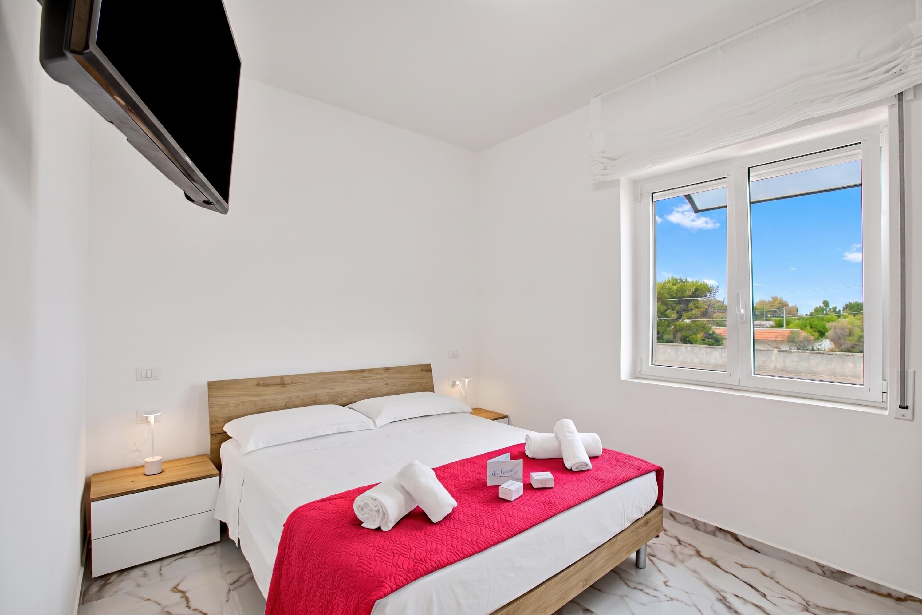 Quarto de hotel com cama, mesa de cabeceira, televisão montada na parede e janela com vista para o verde.