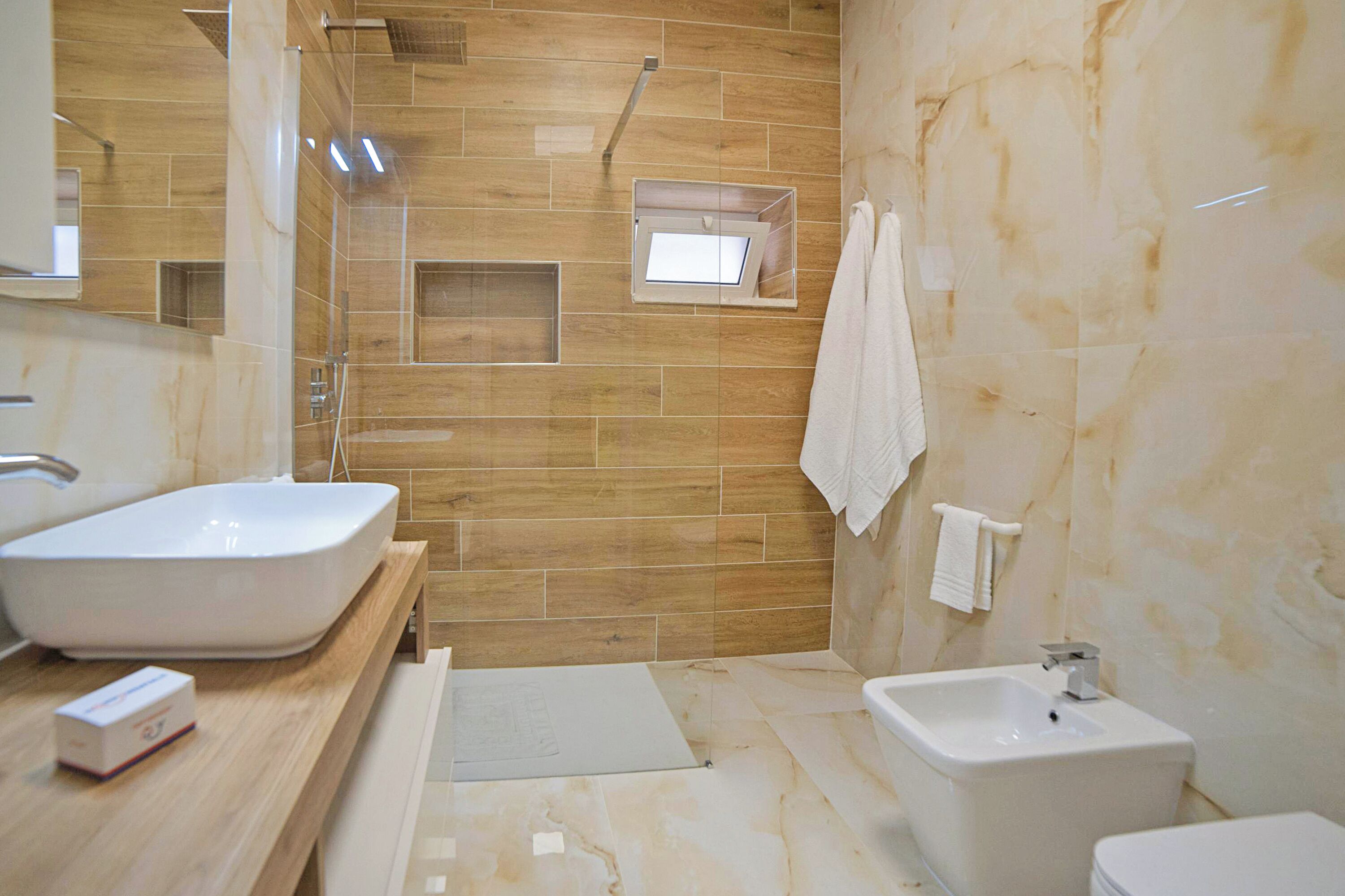A modern bathroom with a large white sink, a bidet, and a toilet. There is a wooden vanity with a mirror and a small shelf.