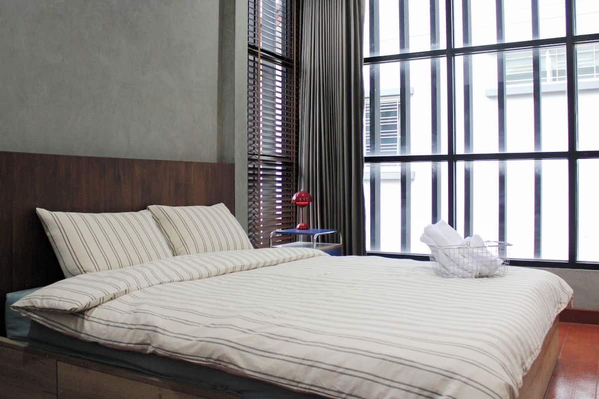 A neatly made bed with striped bedding, a bedside table with a lamp, and a window with blinds.