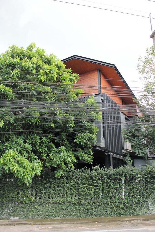 A modern building with a sloped roof, surrounded by dense greenery and a metal fence. - The Melatonin Hostel  (Hat Yai)
