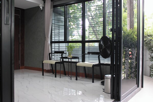 A room with a large window, a fan, a trash bin, and a table with chairs. - The Melatonin Hostel (Hat Yai)