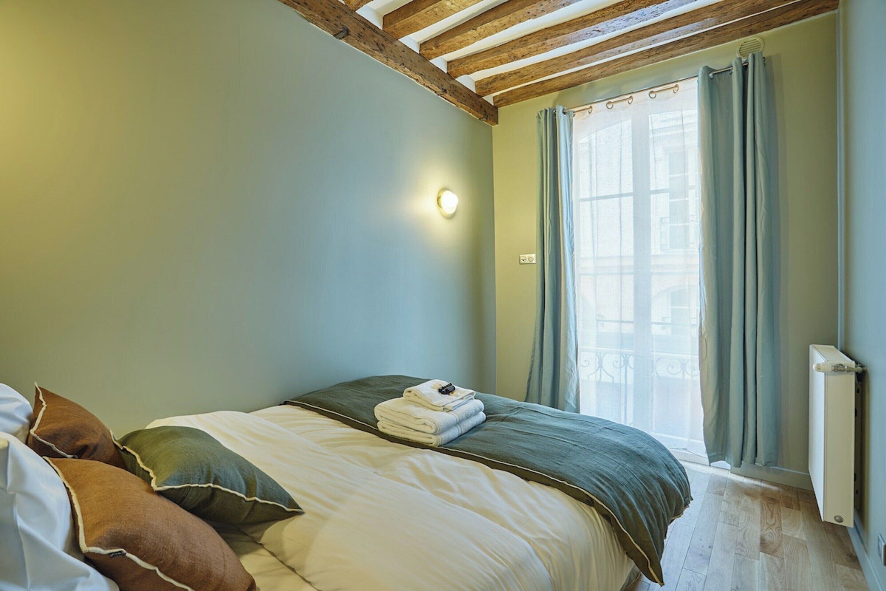Une chambre à coucher avec un lit, des oreillers, un radiateur et une fenêtre avec des rideaux.