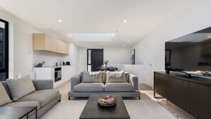 A modern living room with a grey sofa set, a coffee table with fruit, and a kitchen in the background.